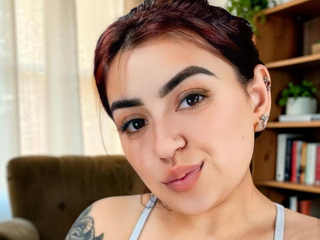 Mariana Rusell a les cheveux teints en rouge, des sourcils foncés, un petit nez et un tatouage sur son épaule gauche. Elle sourit avec un maquillage léger, porte un haut gris et a des piercings aux oreilles..