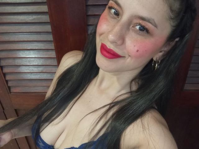 MadissonFontaine tem cabelos pretos longos, batom vermelho e um sutiã de renda azul, mostrando seus seios, com bochechas rosadas e uma pequena pinta na bochecha direita, sorrindo para a câmera..