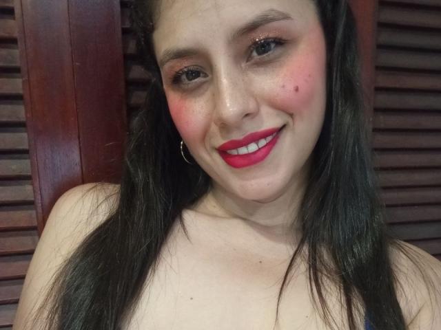 MadissonFontaine tem um leve rubor nas bochechas, batom vermelho e uma pequena espinha no nariz. Ela está sorrindo, usando brincos grandes e tem cabelos longos e pretos..