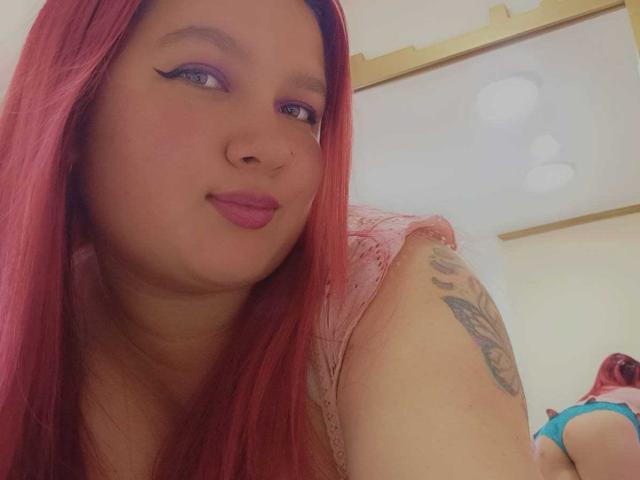 SarahCoupper with pink hair, purple eyeliner, pink lips, tattoo on arm, wearing pink top, smiling, lying down, blue underwear, visible ass, soft lighting, casual selfie.