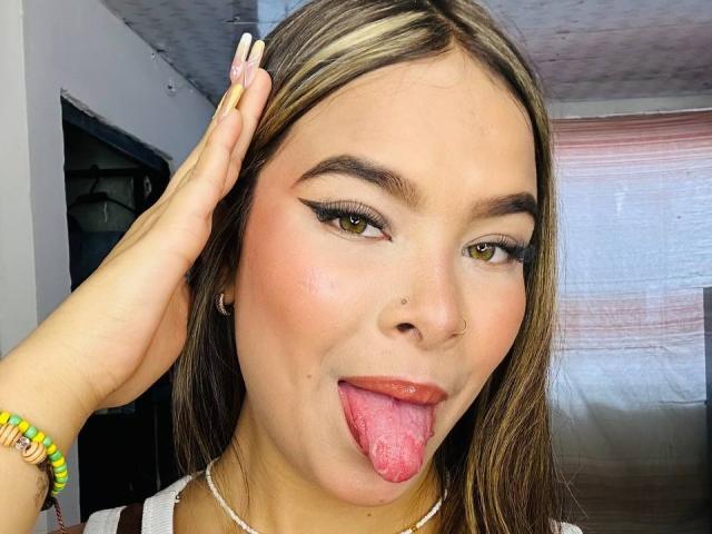 AdharaCopper with blonde highlights, green eyes, and nose ring, sticking out her pink tongue, hand on head, wearing a white shirt and colorful bracelet.