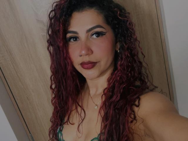 Antonella Rivera, com cabelo ruivo-rosado cacheado, sobrancelhas escuras, delineador de olhos, piercing no nariz, batom escuro, vestido verde, seios médios, pele clara, sorriso discreto, fundo com porta de madeira..