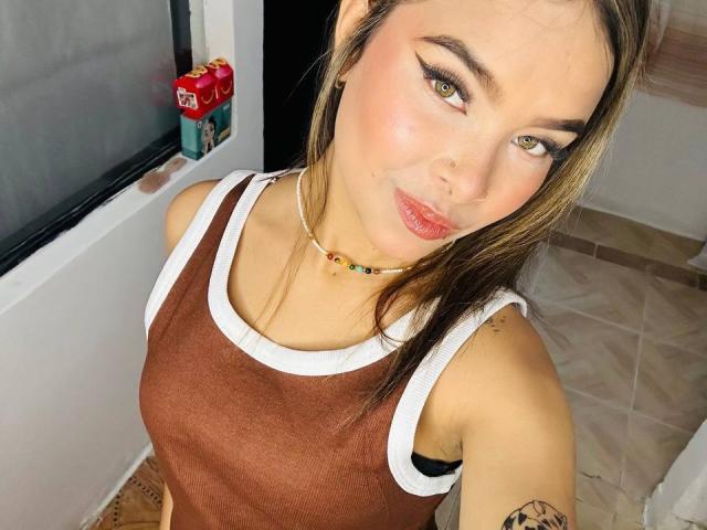 AdharaCopper with light brown tank top, white trim, and colorful bead necklace, has green eyes, pink lips, and brown hair, showing tattoo on right arm, in a tiled room.