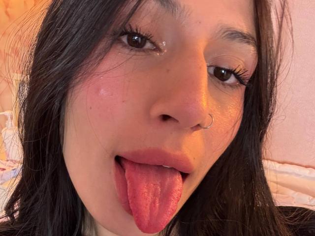 SophieBeaumont with dark hair, brown eyes, and a nose ring, sticking out her pink tongue. She has glossy eyelashes and a slight blush on her cheeks.