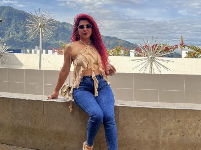 Antonella Rivera mit roten Haaren, die eine Sonnenbrille trägt, ein geripptes, beiges Oberteil und blaue Jeans, sitzt auf einer Betonmauer und zeigt ihr Gesäß in engen Jeans, draußen mit Bergen im Hintergrund..