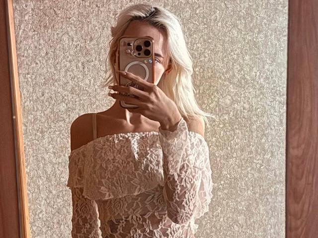 DortheyRosenbluth loira, com cabelo ondulado, usando uma blusa de renda com um ombro descoberto, segurando um celular, cobrindo o rosto, tirando uma selfie no espelho, mostrando os seios, em luz suave, moldura de espelho de madeira..
