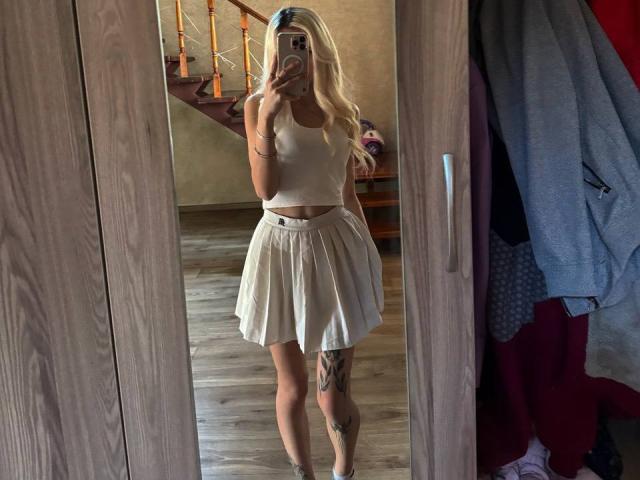Blonde Dorthey Rosenbluth in einem weißen Crop-Top und einem Faltenrock, hält ein Telefon, zeigt Tattoos am Bein, steht vor einem Spiegel, Holzboden, Treppe im Hintergrund..