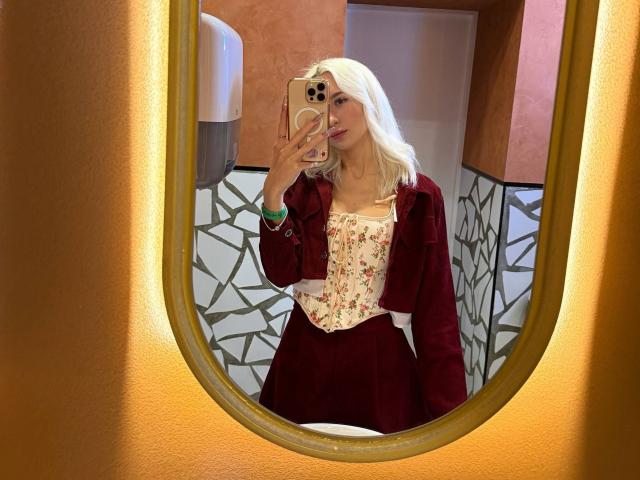 Dorthey Rosenbluth, avec ses cheveux blonds, portant une veste en velours rouge et un haut blanc à motifs floraux, prend un selfie devant un miroir, montrant ses seins dans le reflet, tout en tenant un iPhone, dans une salle de bain..