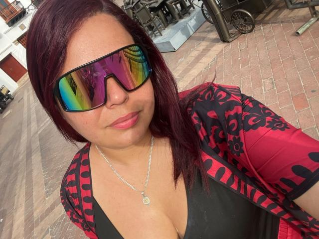 Maya Malik, avec ses cheveux roux, portant des lunettes de soleil réfléchissantes, une chemise noire et rouge à motifs, et un collier en argent, montrant sa poitrine généreuse dans un haut noir, se tient dans une zone bordée de briques..