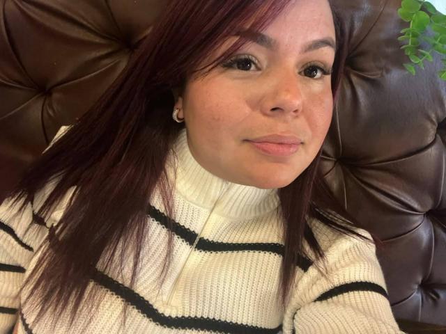 MayaMalik has a cute face with medium brown skin, straight red hair, and is wearing a white striped turtleneck, sitting on a brown leather couch.