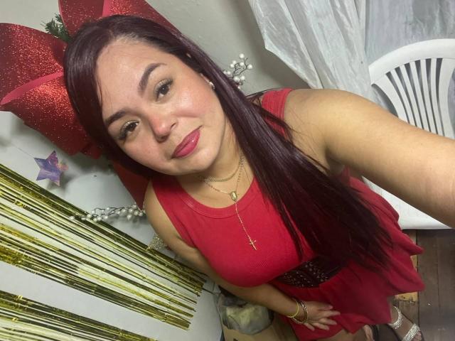 Maya Malik, avec de longs cheveux violets, une robe rouge et un collier avec une croix, debout, regardant la caméra, souriant, un nœud rouge, une étoile dorée et des décorations en bambou en arrière-plan, les mains sur la robe..
