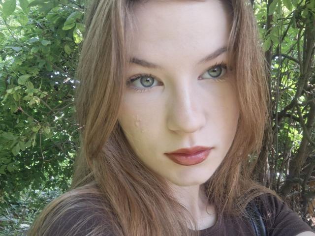 Justine Hasenfratz, met groene ogen, lichtbruin haar en rode lippenstift, draagt een bruin shirt en heeft een traan op haar rechterwang. Ze kijkt recht in de camera..
