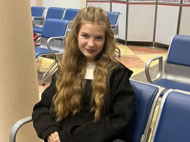 Justine Hasenfratz, mit langen, welligen blonden Haaren und einer schwarzen Kapuzenjacke, sitzt auf blauen Metallstühlen, die Arme verschränkt, und lächelt leicht in einem tristen Wartezimmer..