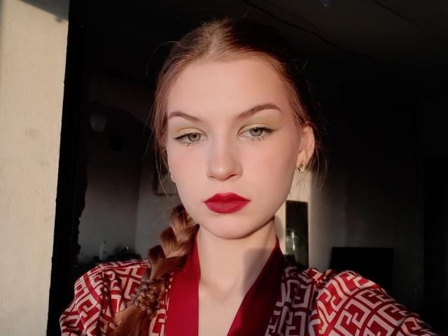 JustineHasenfratz has pale skin, red lipstick, green eyes, and a braid, wearing a red and white patterned shirt, standing in sunlight with a serious expression.