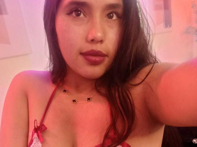 GemmaBellini with long brown hair, red lipstick, wearing a red halter top, necklace with small black star pendants, looking at the camera, pink-red light. Boobs visible.