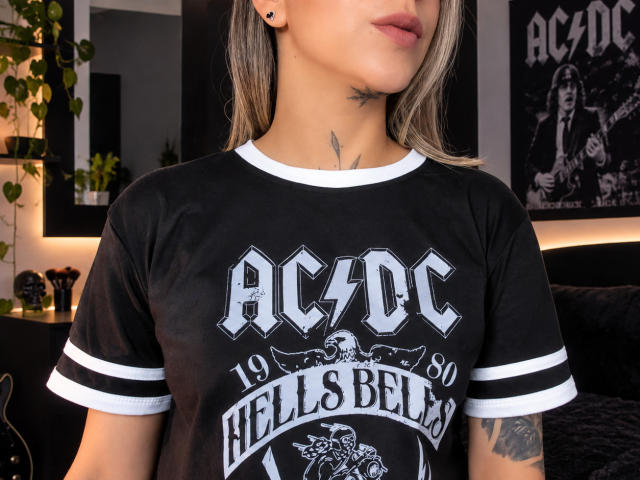 KathyM, avec des cheveux blonds, un t-shirt noir AC/DC, des finitions blanches, de petits tatouages sur le cou et le bras, les lèvres légèrement entrouvertes, des piercings aux oreilles, dans une pièce faiblement éclairée..
