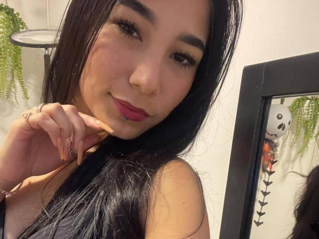 VioletaVelvet with long black hair, brown eyes, and pink lipstick, smiling at the camera, holding her hair with one hand, showing off her golden nails and ring.