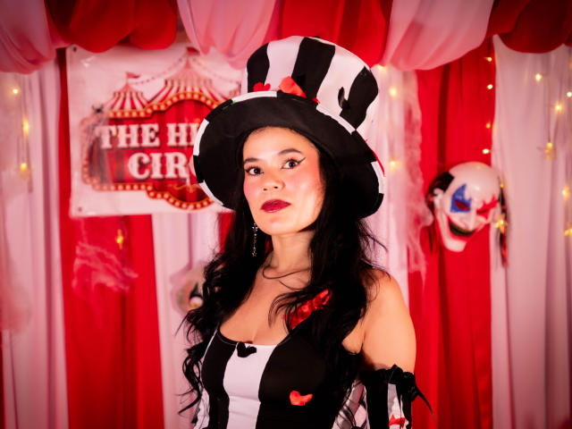 Isabella Voss usando um chapéu listrado preto e branco, vestindo um vestido preto e branco com detalhes vermelhos, com cabelo preto longo, batom vermelho e brincos vermelhos, olhando para a câmera..