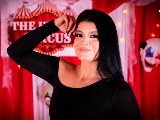 VictoriaFiore with long black hair, wearing a black long-sleeve top, smiling slightly, hand near head, red curtains and string lights in background, looking sexy and confident.