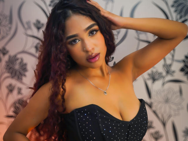 MadisonDawson with curly dark red hair, wearing a black strapless dress with small white dots, her hand in her hair, a silver necklace, and purple lipstick. Her tits are visible.
