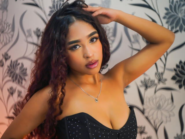 Madison Dawson, com cabelo ruivo-acastanhado encaracolado, pele marrom média e um piercing no nariz, vestindo um vestido preto sem alças com brilhos, com a mão no cabelo, olhando para a câmera, usando um colar, com maquiagem suave..