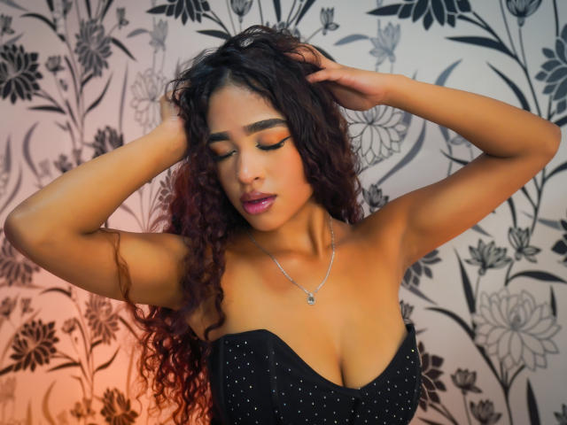 MadisonDawson with curly red-black hair, eyes closed, wearing a strapless black dress with sparkles, hands in hair, necklace, and pink lipstick, showing her tits.