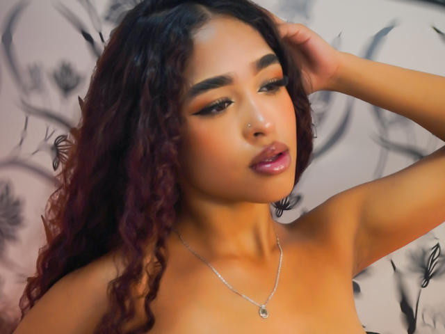 Madison Dawson, com cabelos ruivo-acastanhados cacheados, sobrancelhas escuras, delineador e lábios rosados brilhantes, usando um colar de prata e um piercing no nariz, com uma mão no cabelo..