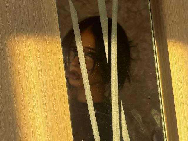 ShanekaKonig's face is partially visible through a window with vertical white lines, her dark hair and glasses slightly blurred by the glass. Her mouth is slightly open.