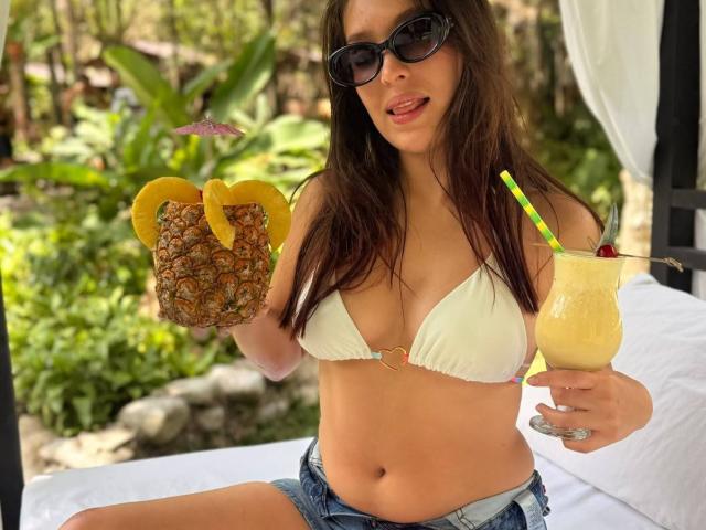Katarina Novak mit braunen Haaren, die schwarze Sonnenbrille trägt und ein weißes Bikini-Oberteil anhat, hält ein Getränk mit Ananas und Zitronenscheiben sowie einen gelben Cocktail mit einem grünen Strohhalm..