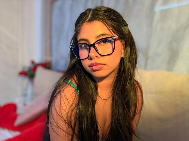 Celeste Morel, met lang bruin haar, een zwarte bril, een groene band, kijkt naar de camera, heeft volle lippen, subtiele make-up, warm licht, zachte focus, een subtiele glimlach, is schattig en aantrekkelijk..