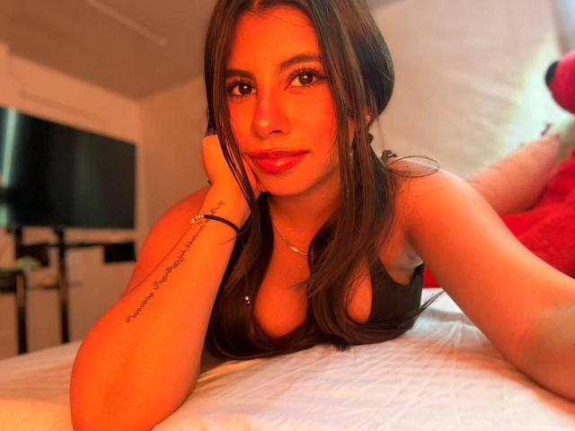 CelesteMorel, with long brown hair and red lipstick, leans on a bed, wearing a black top, showing her tits, with a tattoo on her right arm, in warm light.