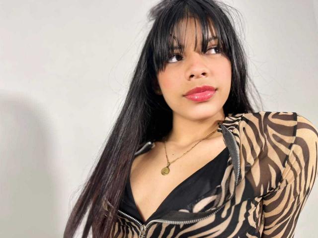 JeanneLacroix has long black hair, bangs, and a zebra-patterned top with a black bra visible. She wears a gold necklace and has pink lipstick.