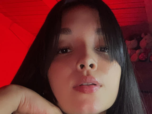 SarahGood's face with black straight hair, glossy pink lips, and a cute nose, looking at the camera with a slight smile, under red lighting, holding her chin.