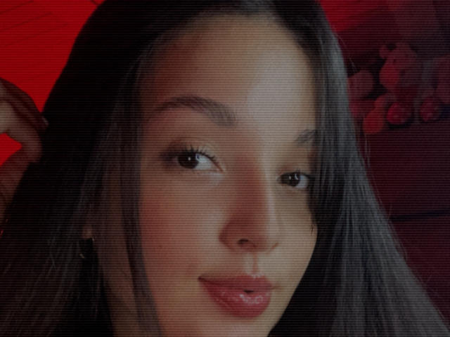 SarahGood has long black hair, brown eyes, and glossy pink lips. She's looking slightly to the side with a subtle smile, and her hand is near her head.