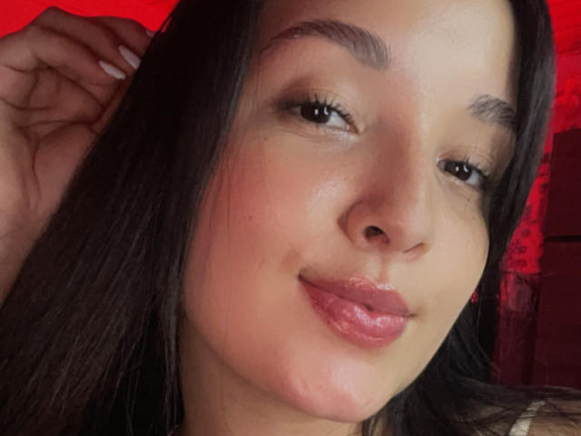 SarahGood's face, with glossy pink lips and dark eyebrows, smiling slightly. She has straight black hair and a fair skin tone. Her right hand touches her head.