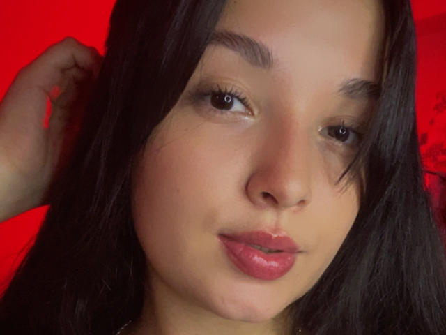 SarahGood's face with medium brown skin, black eyebrows, brown eyes, and pink lips. She has black hair, slightly parted, and is looking at the camera with a neutral expression.
