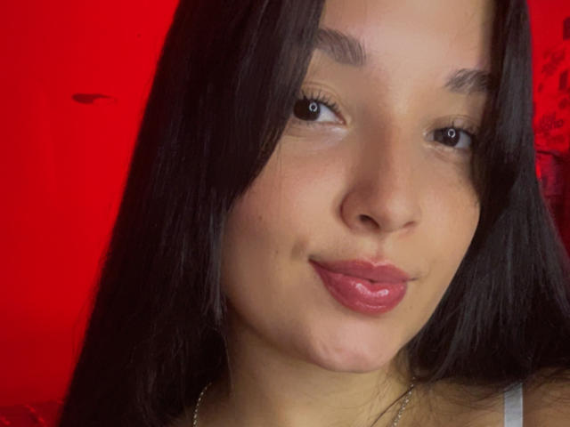 SarahGood has brown eyes, straight black hair, and glossy pink lips with a slight pout. She's looking at the camera with a subtle smile. Red background.