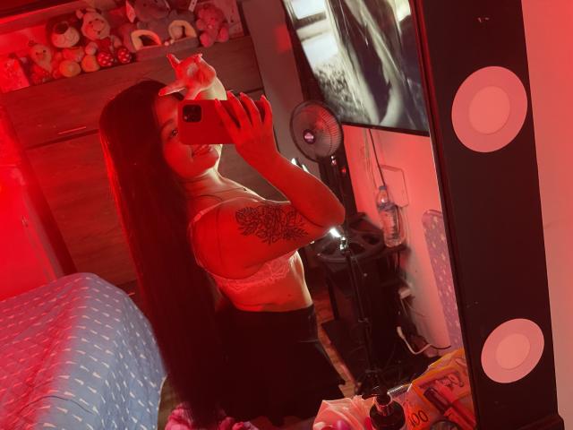 SarahGood with long brown hair, tattoo on right arm, holding phone, mirror selfie, red light, wearing white lace bra, black skirt, visible boobs, bedroom, stuffed toys in background.