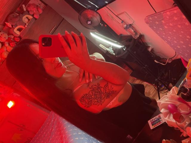 SarahGood with red hair, wearing a black top, showing a large floral tattoo on her right boob, taking a mirror selfie with a red phone in a red-lit room.