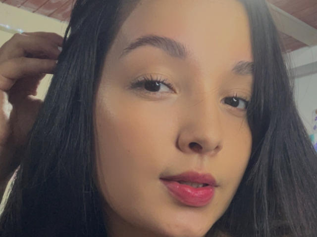 SarahGood's face, with long black hair, brown eyes, and pink lips, slightly parted. She's touching her hair with her left hand. Her skin is light brown.