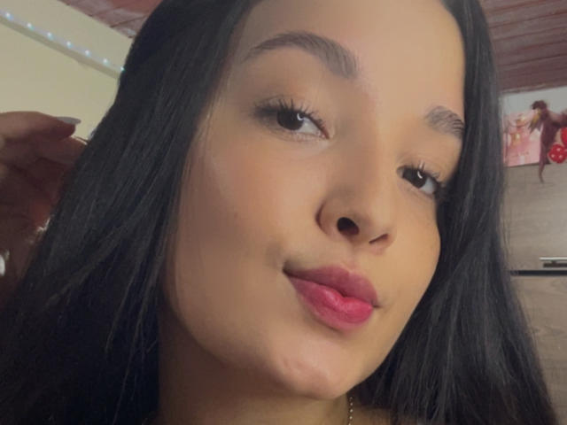 SarahGood's face, brown skin, long black hair, pink lips, slightly raised eyebrows, looking at camera, right hand near head, subtle smile, soft lighting, subtle makeup.