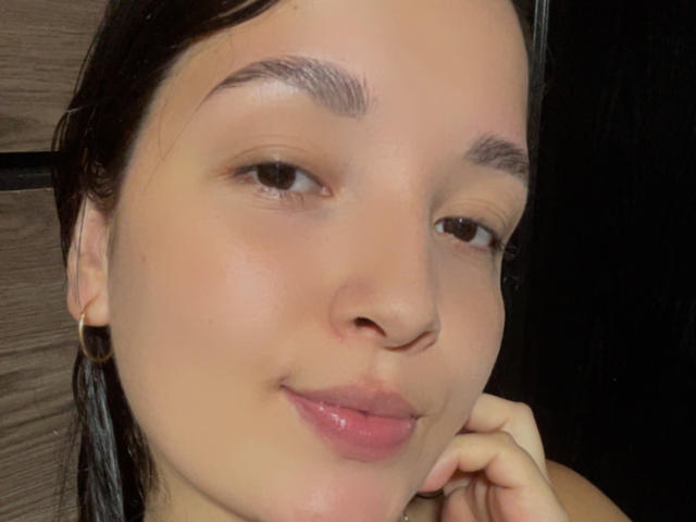 SarahGood's face with light brown skin, brown eyes, small nose, slightly glossy pink lips, dark eyebrows, wet black hair, small gold hoop earrings, and a gentle smile.