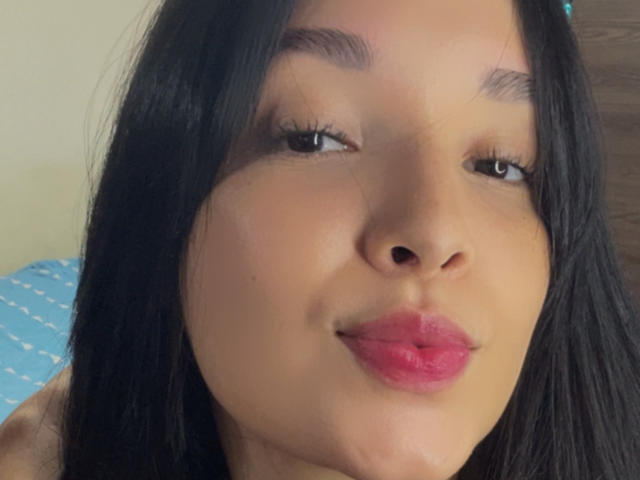 SarahGood's face with brown skin, black hair, and pink lipstick, pouting her full lips. She has dark eyebrows and long eyelashes, looking slightly to the side.