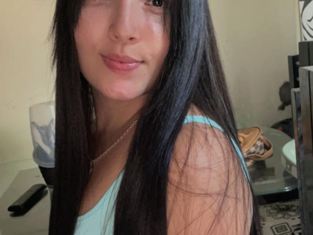 SarahGood with long black hair, light brown skin, wearing a light blue tank top, slight smile, small pinkish lips, small nose, necklace, and a black phone in the background.
