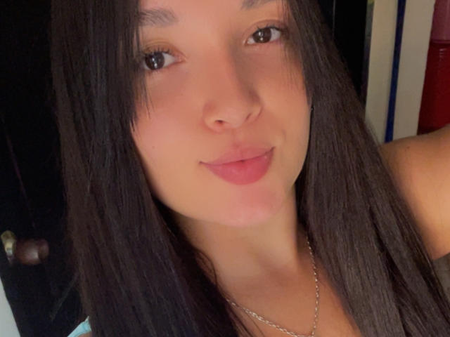 SarahGood has straight dark brown hair, brown eyes, and pink lips. She's wearing a silver necklace and looking at the camera with a slight smile. Her tits aren't visible.