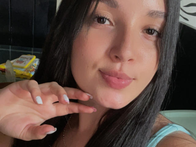 SarahGood has long black hair, light skin, and pink lips, smiling with a slight pout. She's touching her chin with a hand, showing white nails, in a selfie.