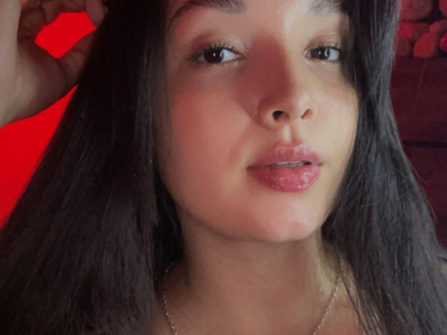 SarahGood has shiny brown eyes, long dark hair, and pink glossy lips. She's wearing a silver necklace with a red background. Her right hand is touching her hair.