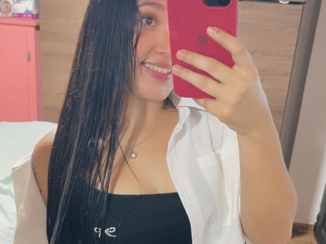 SarahGood with long black hair, smiling, holding a red phone, wearing a white shirt over a black top, showing her tits, in a mirror selfie.