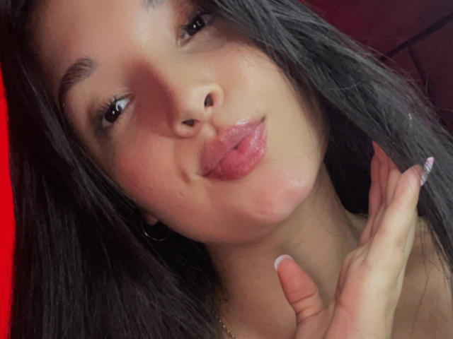 SarahGood with dark hair, slightly pouty pink lips, and her tongue out, holding her neck, wearing hoop earrings, and sporting long, manicured nails.