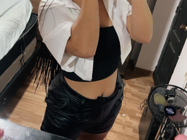 SarahGood's midriff showing a black sports bra and black shiny shorts, with a white shirt partially open, standing in a room with wooden floors and a fan.